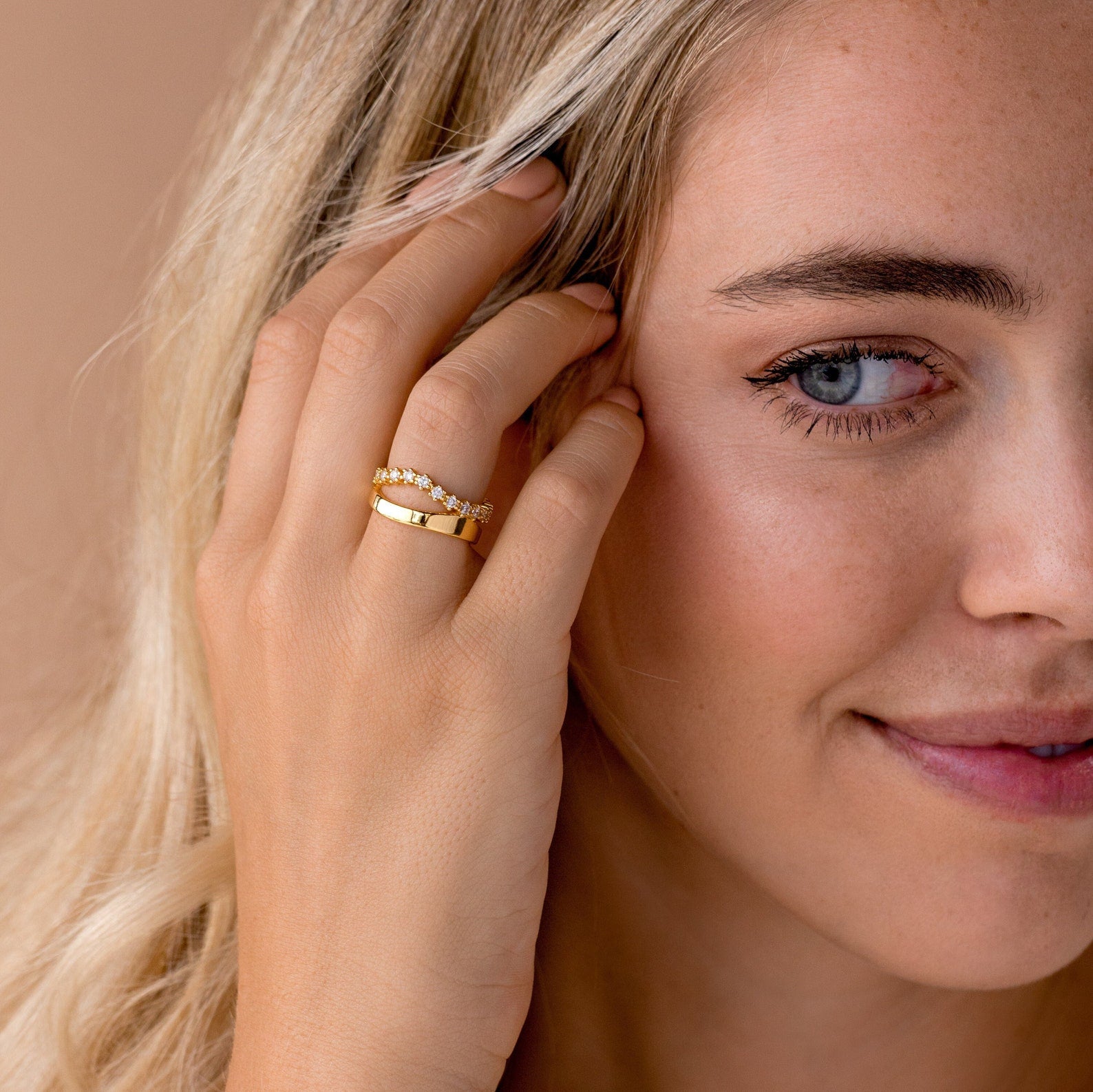 A blonde woman smiles softly, holding her hand near her face to display the Wave Diamond Duo Ring in gold.