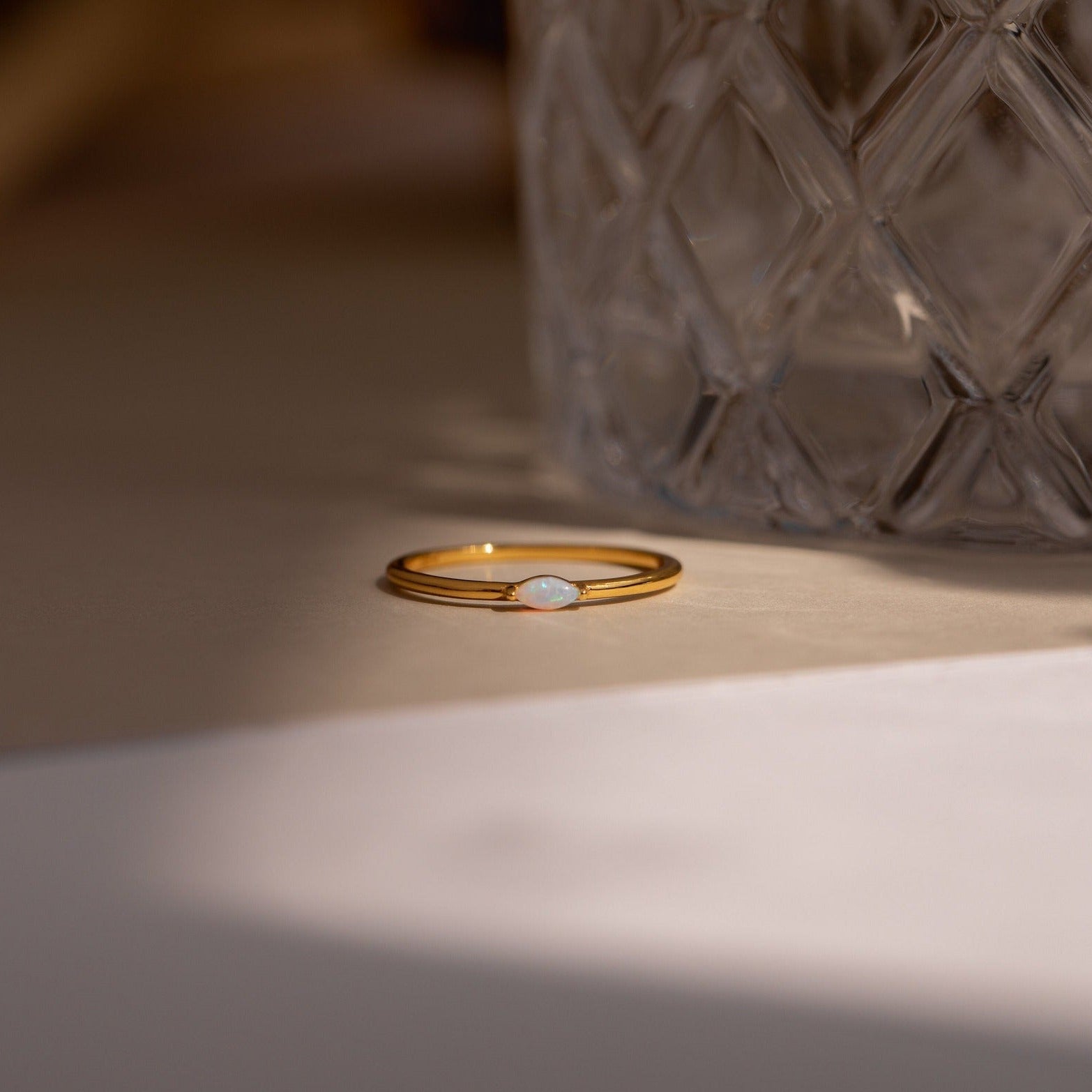 The Eloise Opal Ring, featuring a small iridescent stone, rests beside textured glass in soft lighting, capturing the essence of minimalism.