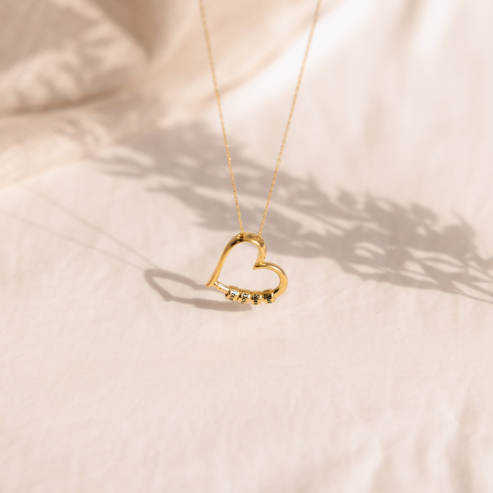 The Custom Bead Heart Necklace, featuring gold and small gemstones, is showcased on a soft, light-colored fabric. Personalized engraved beads make this elegant accessory a timeless and meaningful gift.