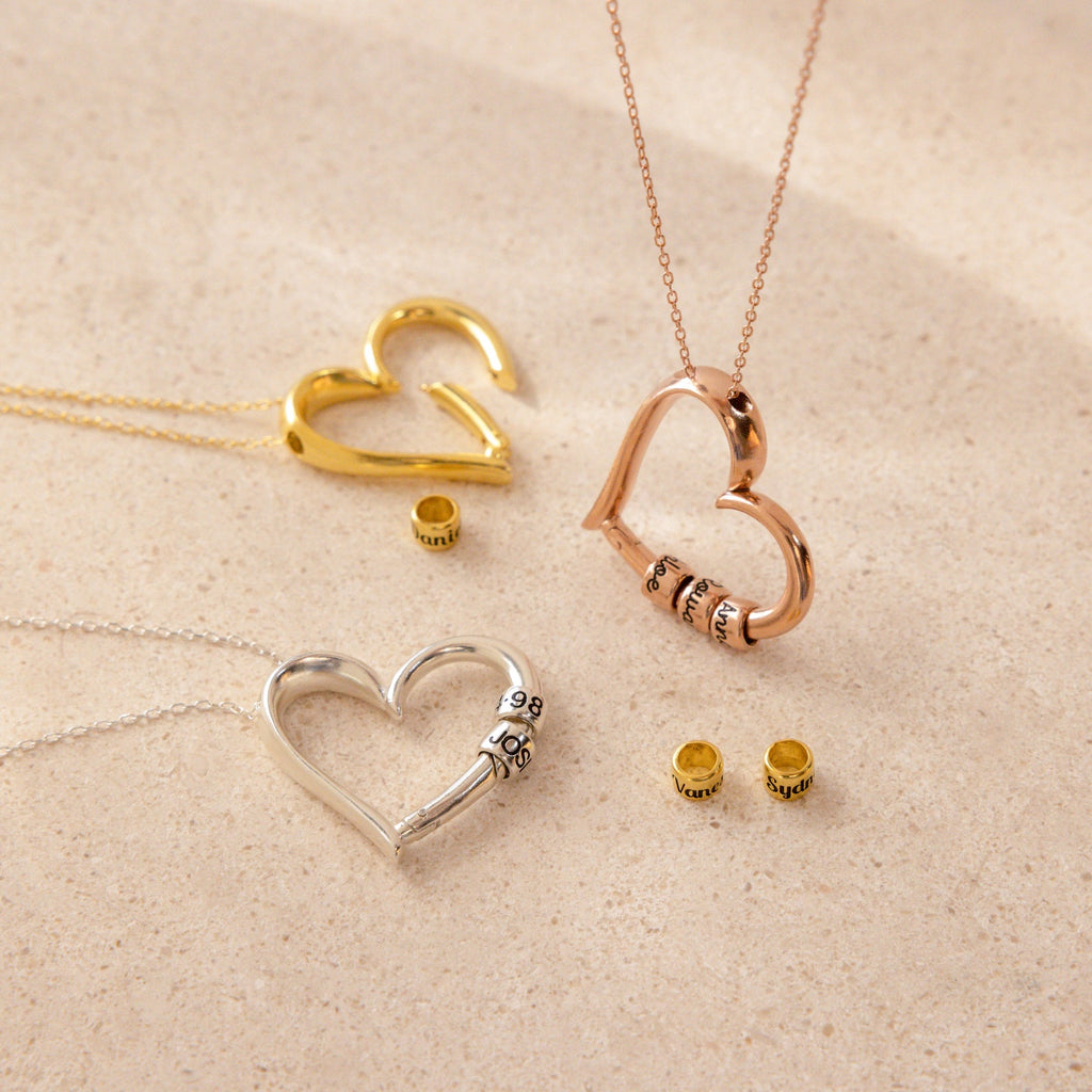Three heart-shaped Custom Bead Heart Necklaces in gold, rose gold, and silver, each with personalized engraved beads, are displayed on a beige surface. Add the perfect custom necklace to your collection today.