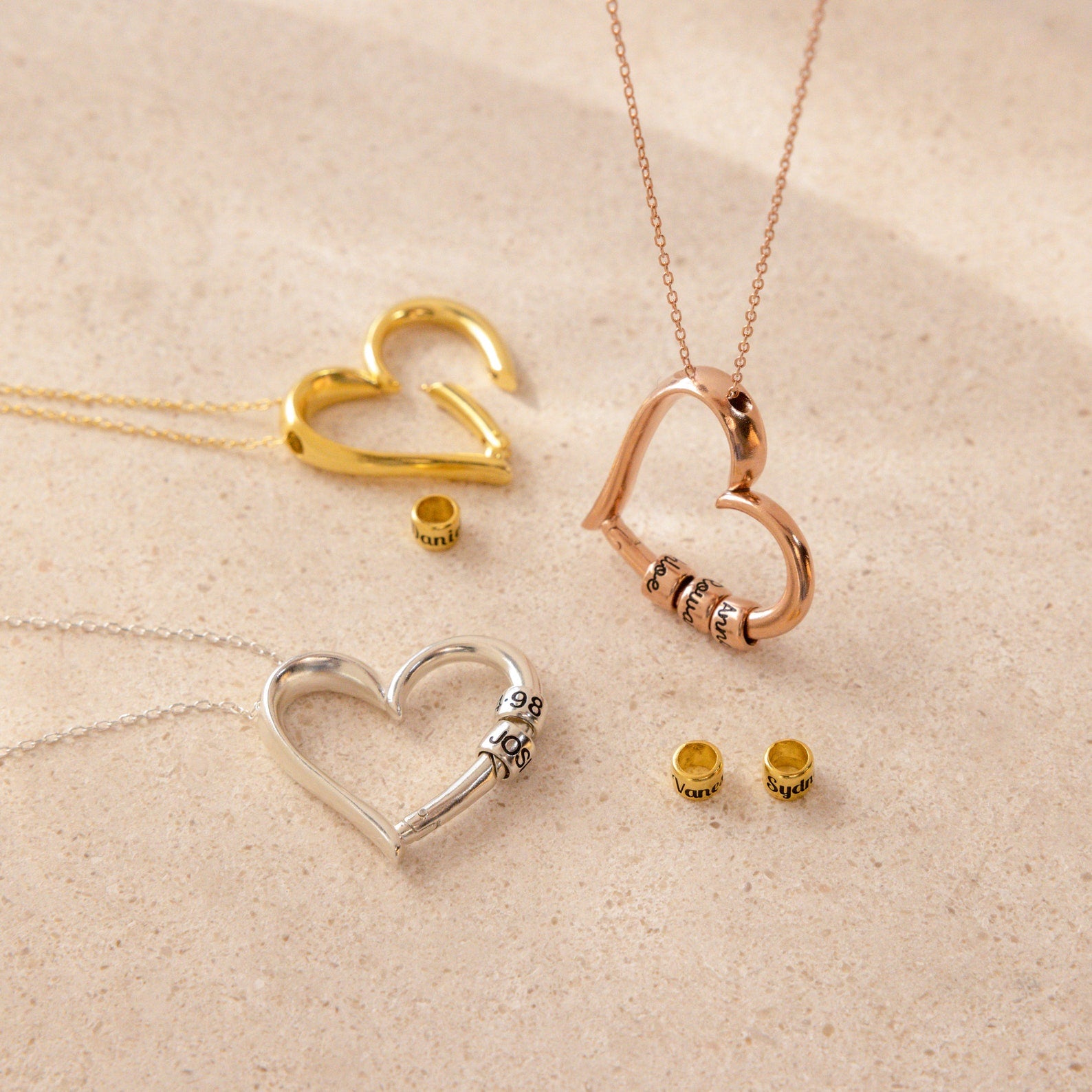 Three heart-shaped Custom Bead Heart Necklaces in gold, rose gold, and silver, each with personalized engraved beads, are displayed on a beige surface. Add the perfect custom necklace to your collection today.