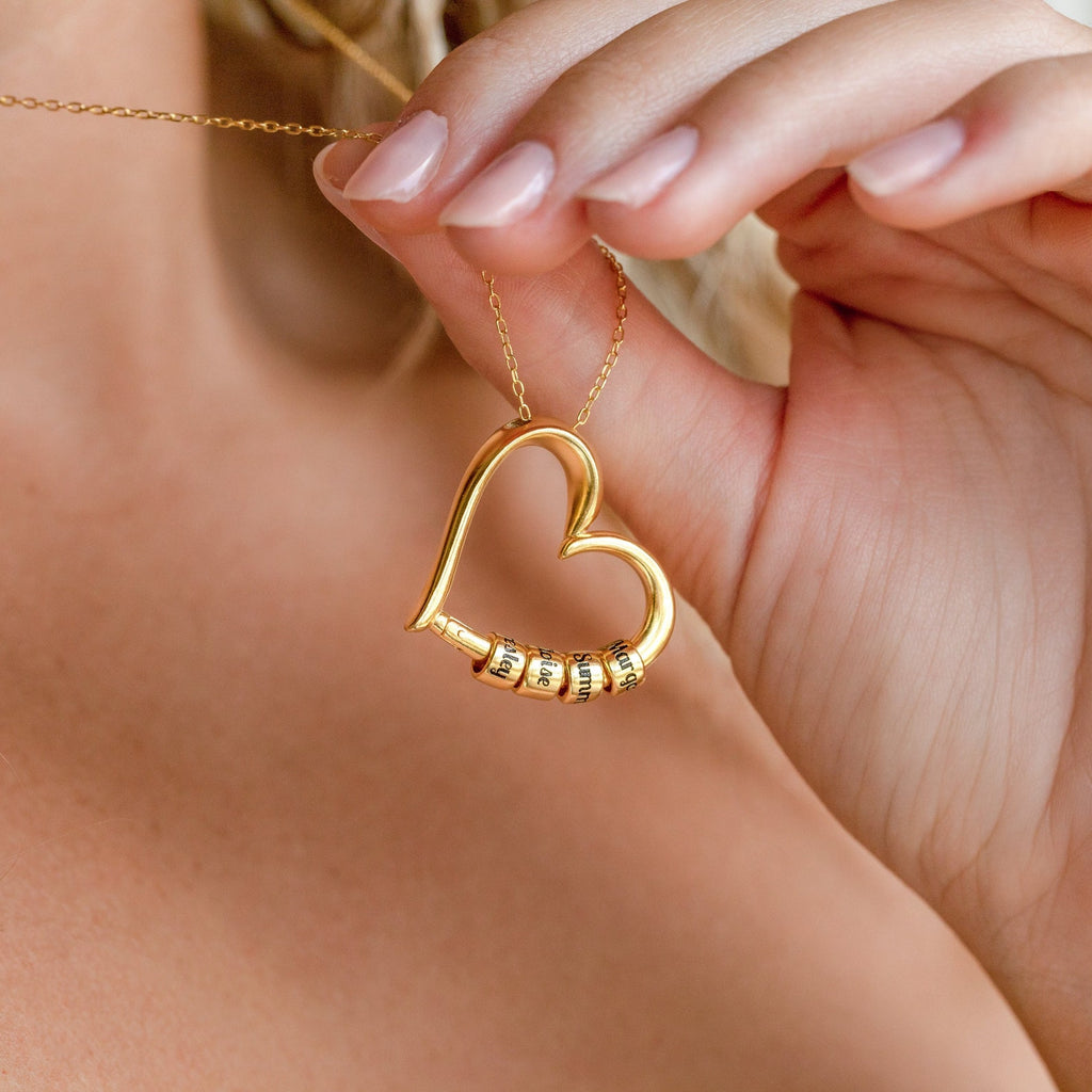 A person holds a Custom Bead Heart Necklace—a gold heart pendant with personalized engraved beads on a delicate chain.