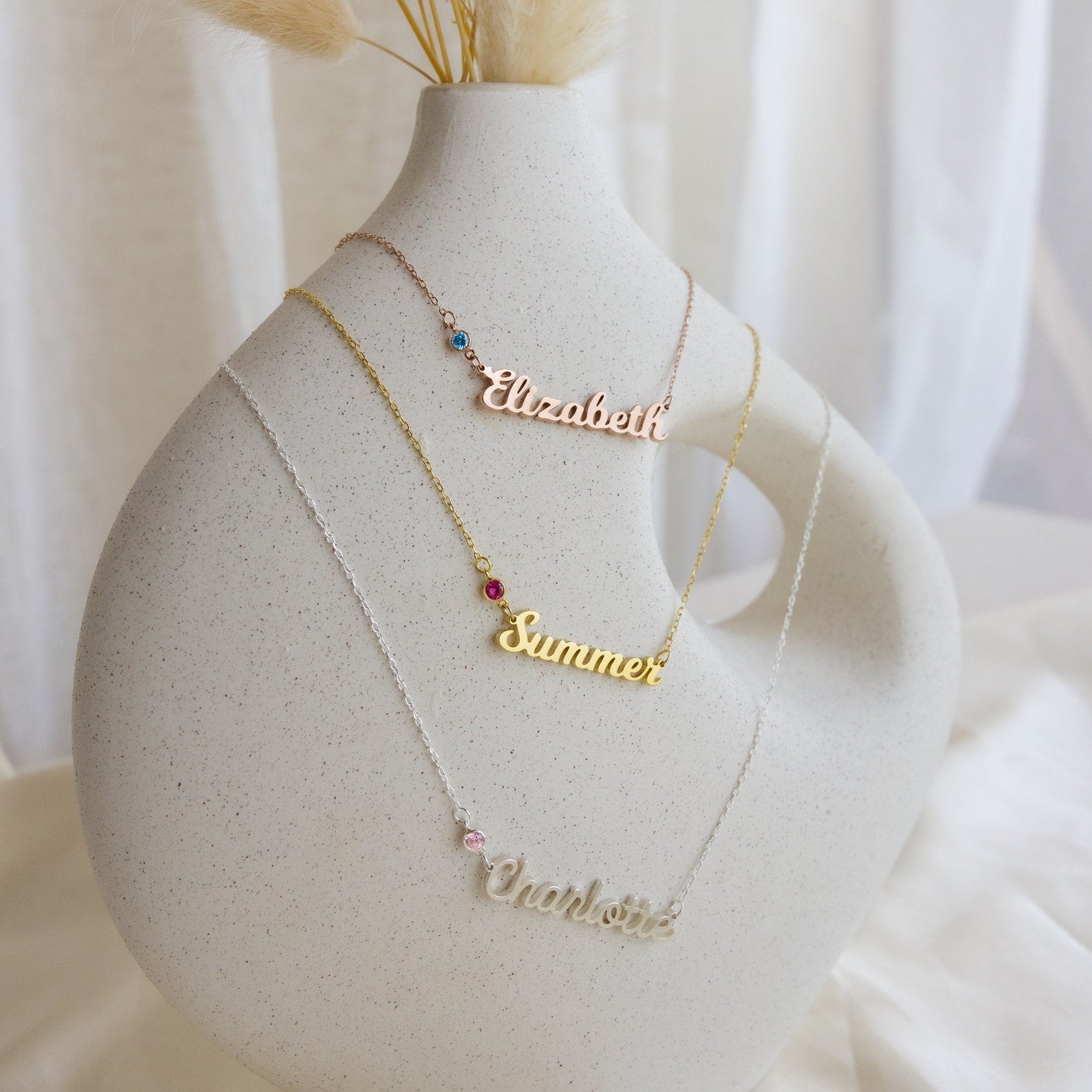 Three Mellow Birthstone Name Necklaces (“Elizabeth,” “Summer,” and “Charlotte”) are displayed on a white ceramic vase, highlighting the elegance of customizable minimalist jewelry—each one made to order for a unique touch.