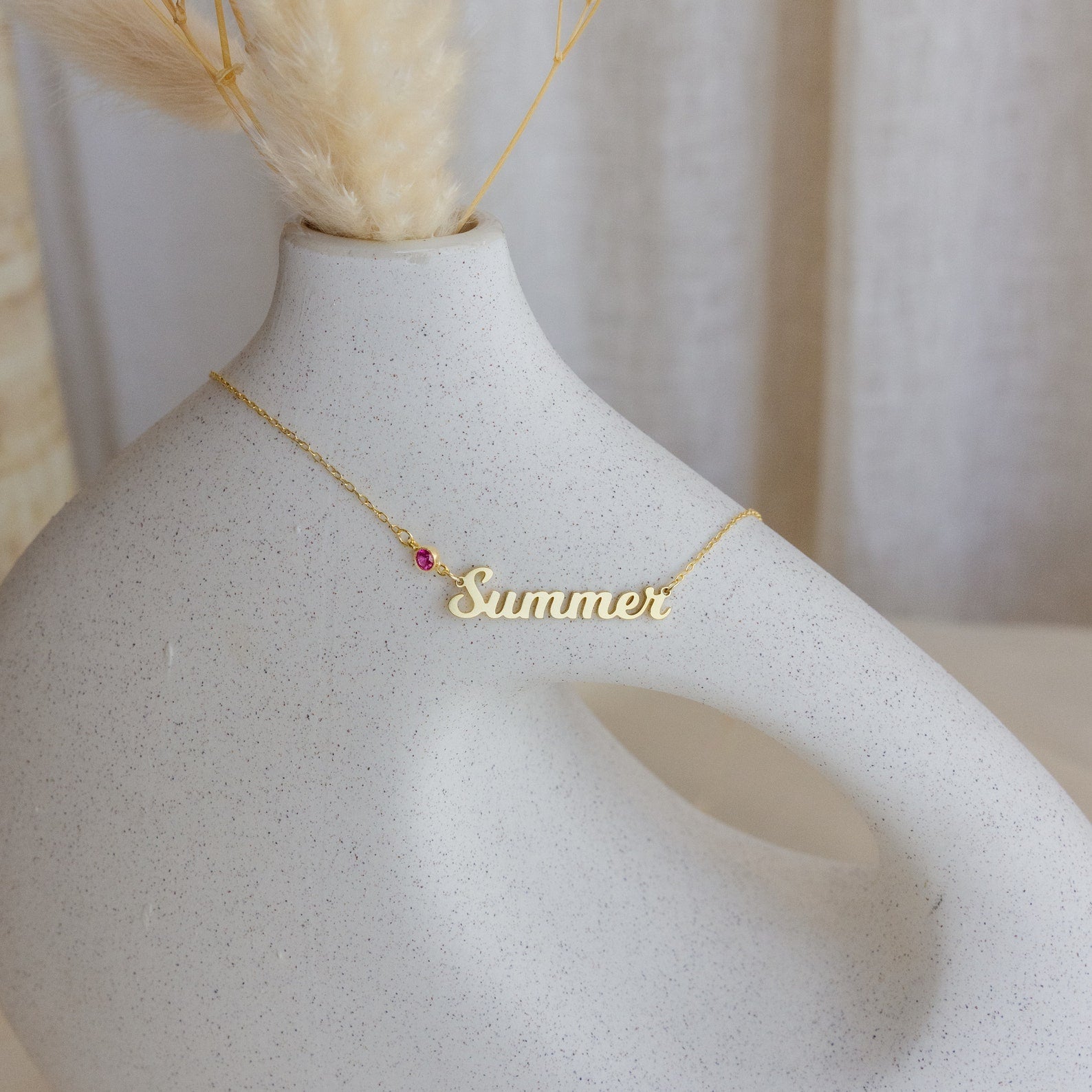 The Mellow Birthstone Name Necklace, featuring a small pink bead, is perfect for minimalist jewelry lovers and is pictured draped over a modern white ceramic vase with dried grasses.