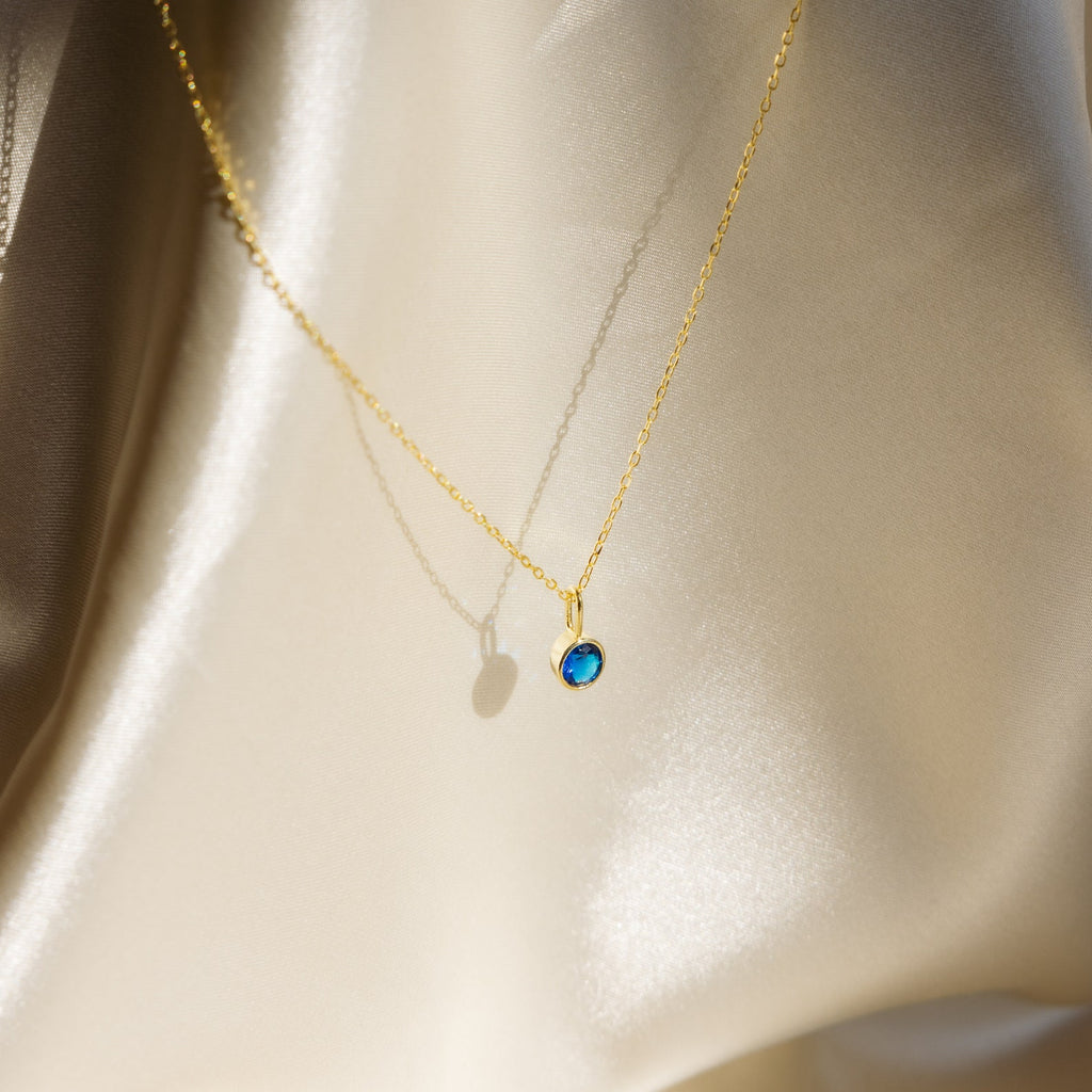 The Custom Birthstone Necklace features a gold dainty chain with a small blue gemstone pendant, making it a meaningful jewelry piece or birthstone necklace, beautifully displayed on silky, cream-colored fabric.