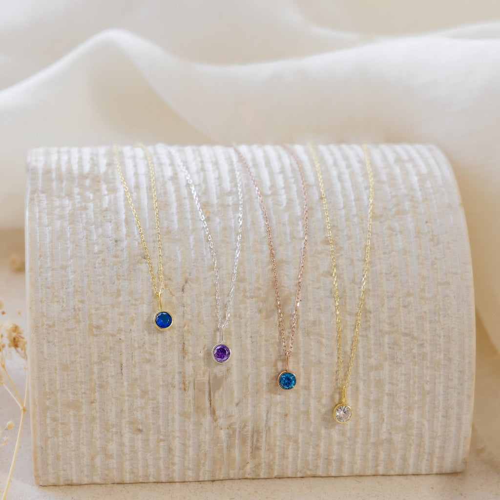 Five dainty Custom Birthstone Necklaces with colored gemstone pendants are displayed on a textured, curved off-white surface—perfect as meaningful jewelry or the ultimate personalized gift.