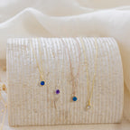 Five dainty Custom Birthstone Necklaces with colored gemstone pendants are displayed on a textured, curved off-white surface—perfect as meaningful jewelry or the ultimate personalized gift.