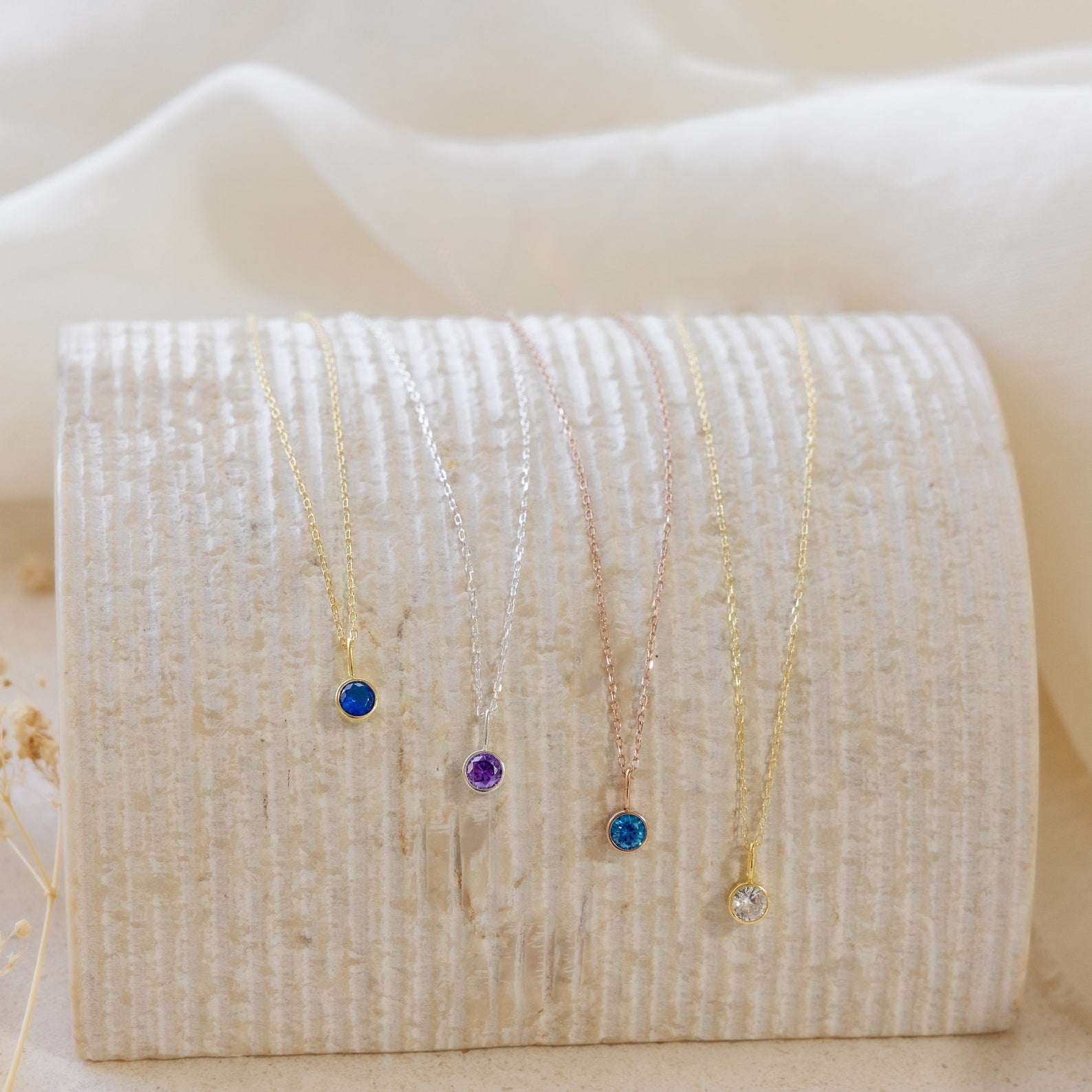 Five dainty Custom Birthstone Necklaces with colored gemstone pendants are displayed on a textured, curved off-white surface—perfect as meaningful jewelry or the ultimate personalized gift.