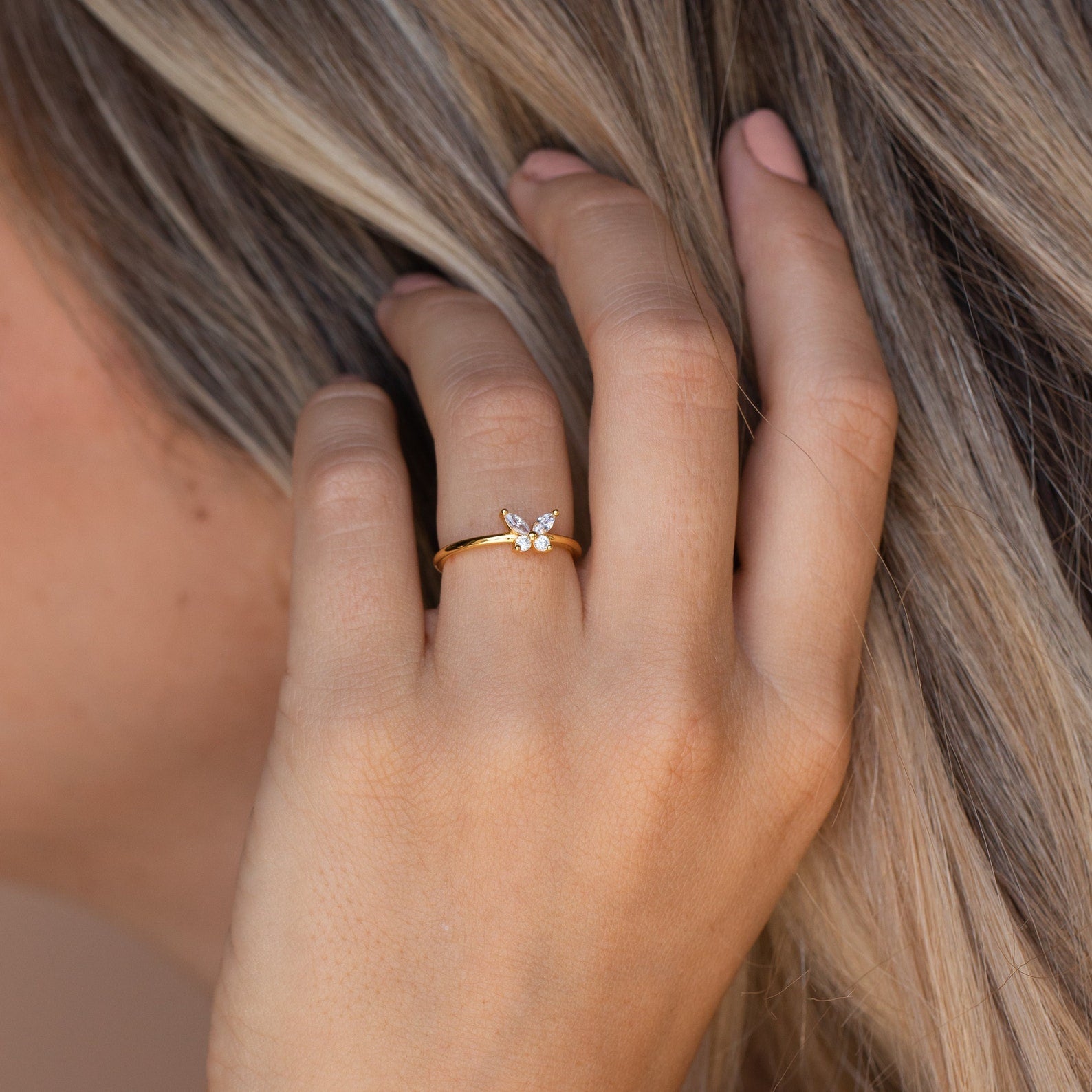 A hand with light nails wears the Mariposa Diamond Ring, a gold butterfly promise ring featuring a delicate floral gemstone design, shown near blonde hair.
