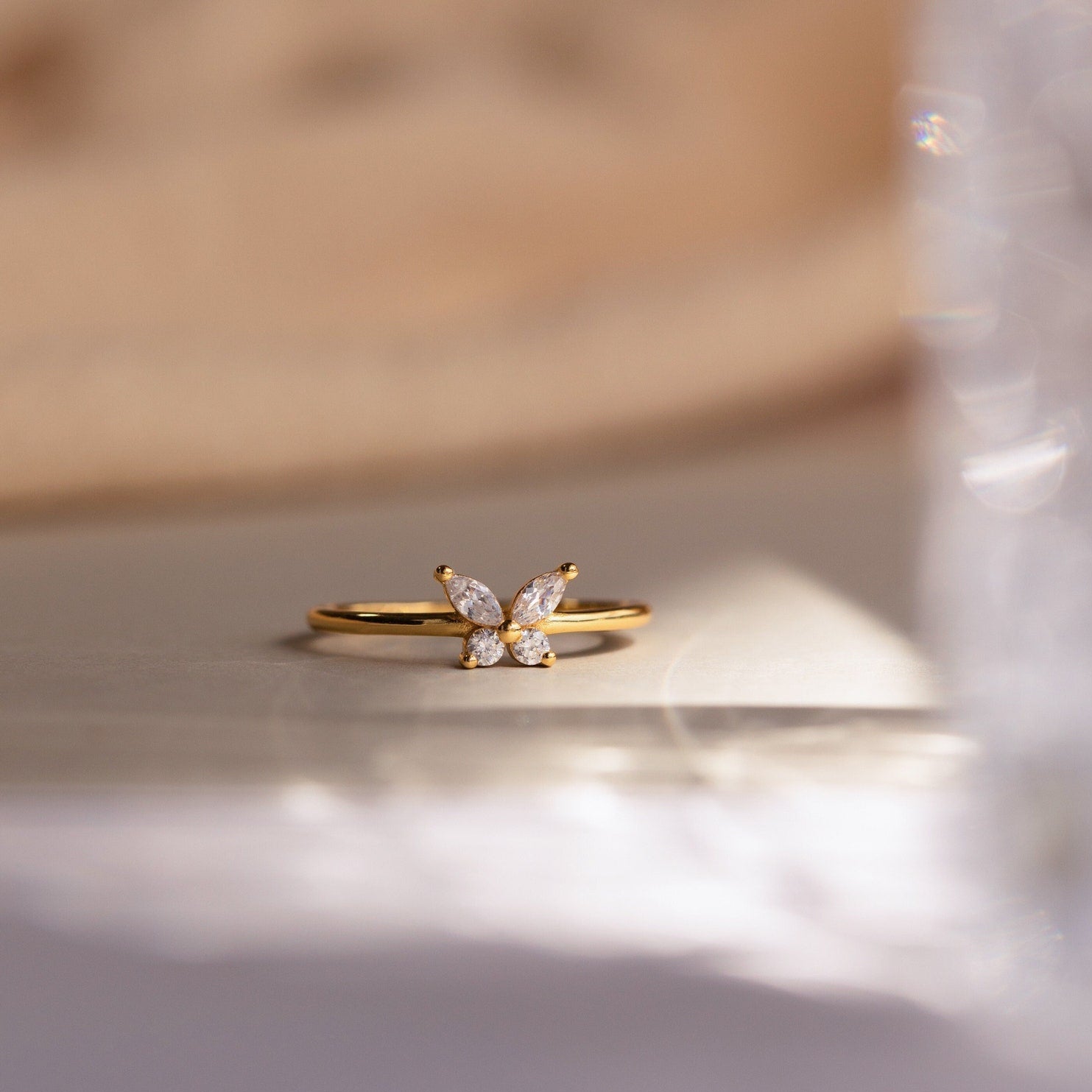 The Mariposa Diamond Ring is a gold butterfly promise ring adorned with a cluster of four sparkling gemstones, beautifully presented on a soft, light surface.