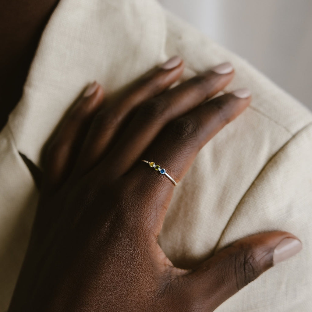 A hand with neatly manicured nails rests on a shoulder, highlighting the Tiny Triple Birthstone Ring—its thin band and trio of tiny gemstones offer a touch of subtle elegance.