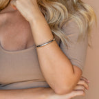 A woman with wavy blonde hair and a beige ribbed top wears the Aris Hinge Bangle, her arm partially raised.