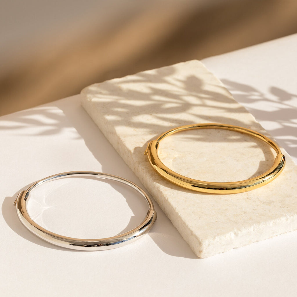 Two Aris Hinge Bangles—one gold, one silver—rest on a white stone surface, highlighted by gentle sunlight and soft shadows.