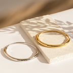 Two Aris Hinge Bangles—one gold, one silver—rest on a white stone surface, highlighted by gentle sunlight and soft shadows.