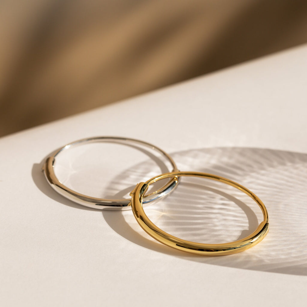 Two Aris Hinge Bangles—one gold, one silver—rest on a white surface with soft shadows, stylishly paired like a modern bracelet stack or minimalist cuff.