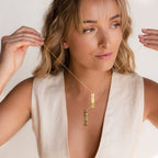 A woman wearing a cream sleeveless top holds and displays two Vertical Handwriting Bar Necklaces, personalized in gold.