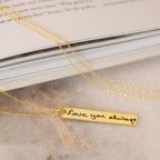 The Vertical Handwriting Bar Necklace is a gold rectangular pendant engraved with "Love you always," shown resting elegantly on a marble surface beside a book.