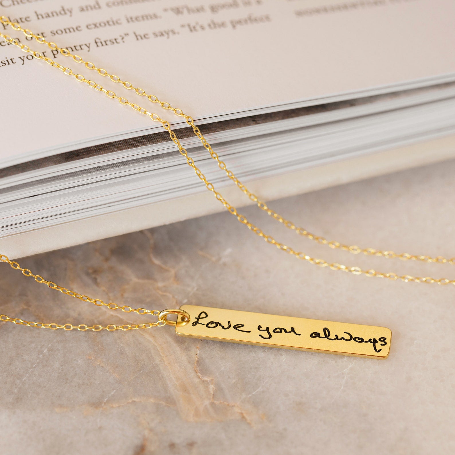 The Vertical Handwriting Bar Necklace is a gold rectangular pendant engraved with "Love you always," shown resting elegantly on a marble surface beside a book.