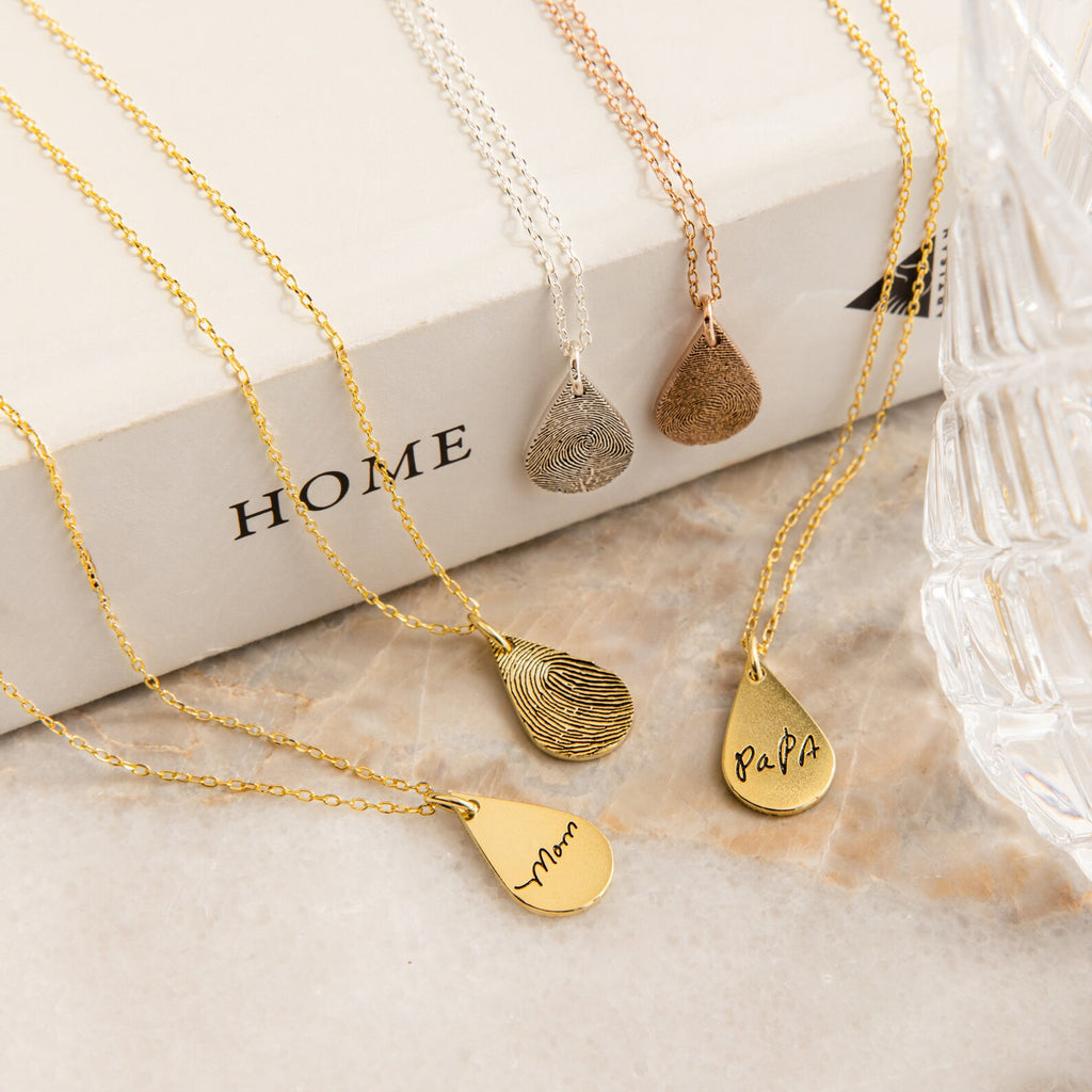 The Tear Drop Fingerprint Necklace, available in gold and silver, is displayed on book and glass surfaces. Some are engraved for personal amulets or as fingerprint keepsakes.