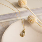 The Tear Drop Fingerprint Necklace rests on a dish beside dried flowers and a closed book—a stunning personalized keepsake featuring your unique fingerprint.