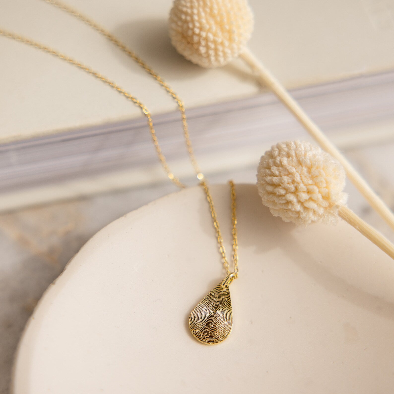 The Tear Drop Fingerprint Necklace rests on a dish beside dried flowers and a closed book—a stunning personalized keepsake featuring your unique fingerprint.