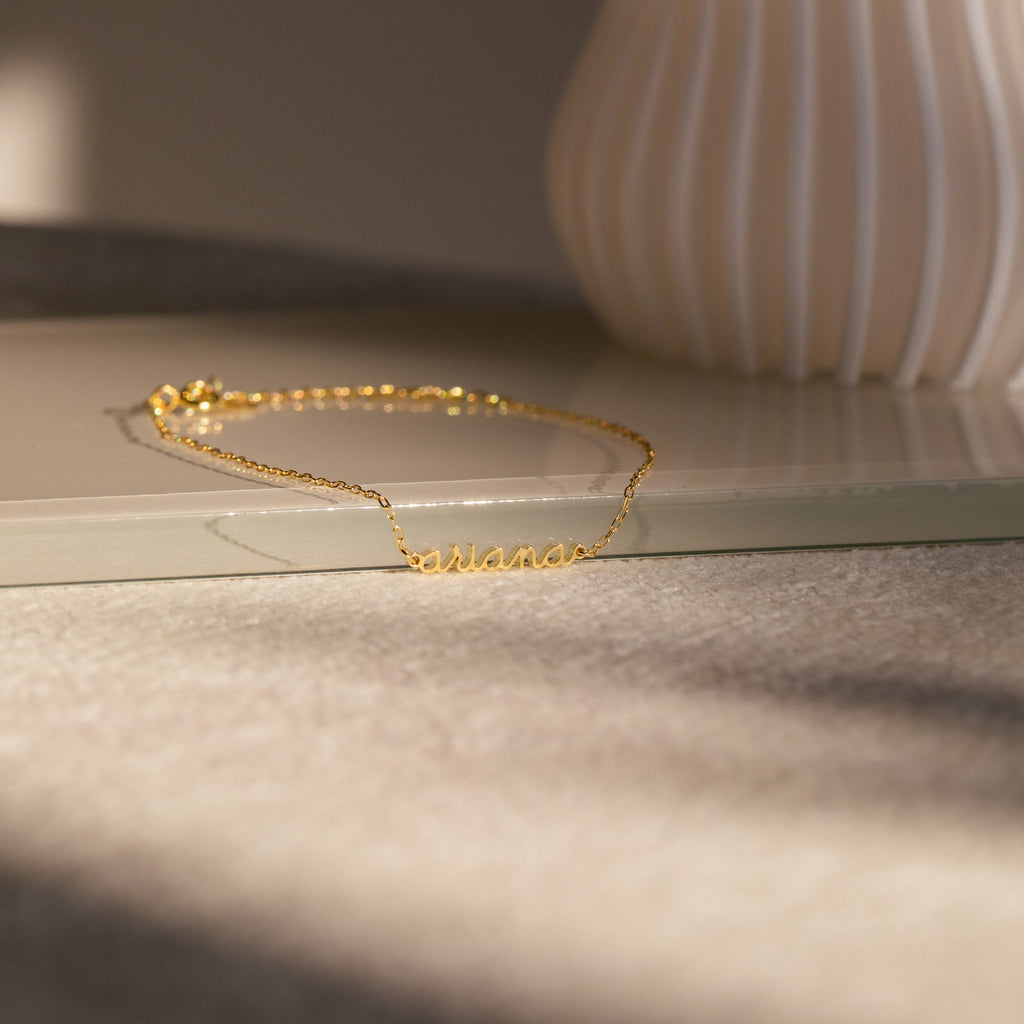 The Dainty Tiny Name Bracelet rests on a beige surface in soft natural light—ideal for fans of minimal jewelry and personalized gifts who love delicate, custom accessories.