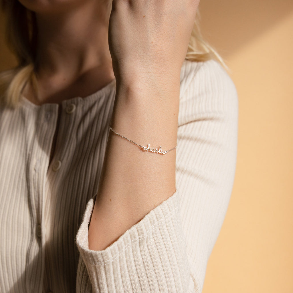 A woman in a cream ribbed top wears the Dainty Tiny Name Bracelet with "Charlie" on her wrist—a perfect piece of minimal jewelry.