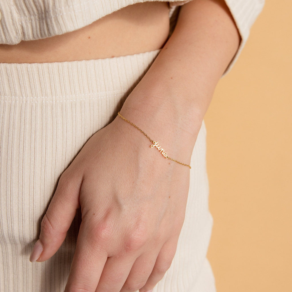 A hand wears the Dainty Tiny Name Bracelet in gold, personalized with "honey," styled as minimal jewelry against a beige background and cream outfit.