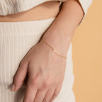 A hand wears the Dainty Tiny Name Bracelet in gold, personalized with "honey," styled as minimal jewelry against a beige background and cream outfit.