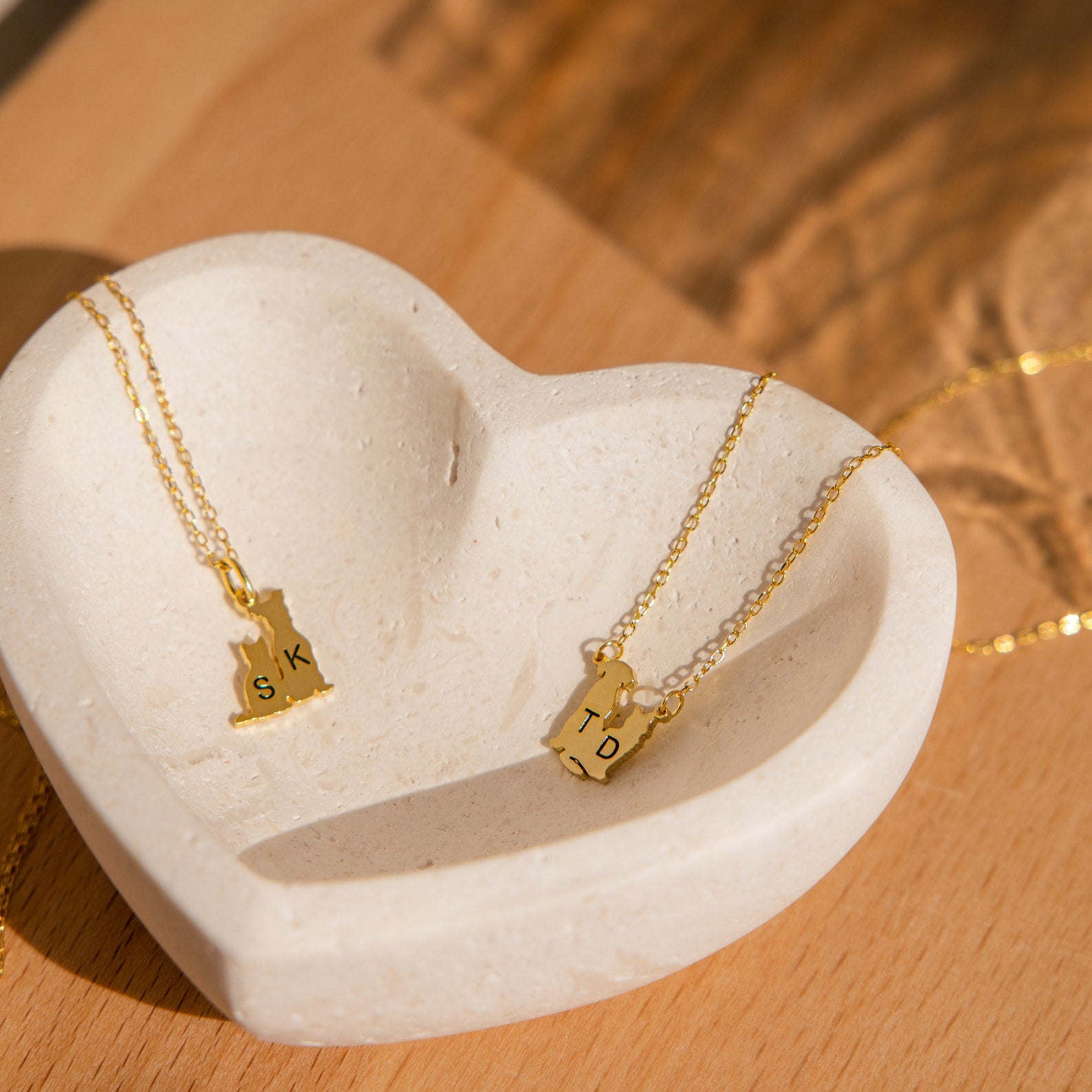 Two Pet Initial Necklaces with state-shaped letter pendants rest in a heart dish on wood, making a perfect personalized gift for pet lovers or anyone who cherishes their furry friends.