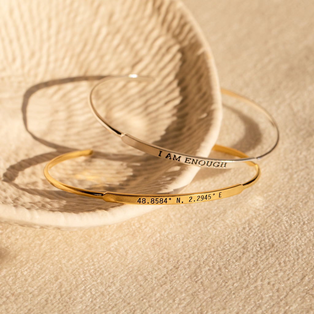 Two Custom Message Cuffs rest on a textured dish; one engraved with "I AM ENOUGH," the other featuring coordinates—a thoughtful, personalized gift.