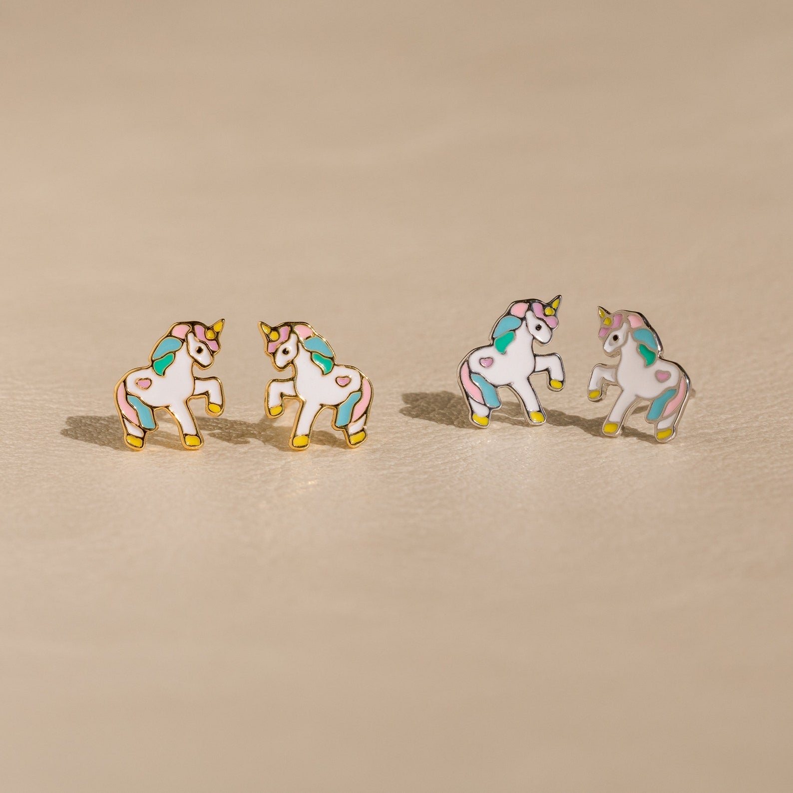 Unicorn Studs: Four pastel enamel unicorn earrings with gold edges on a beige surface—an adorable gift for girls who love whimsical jewelry.