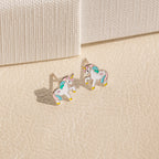 Unicorn Studs with pastel enamel accents rest on a beige textured surface—whimsical earrings and a charming gift for girls.