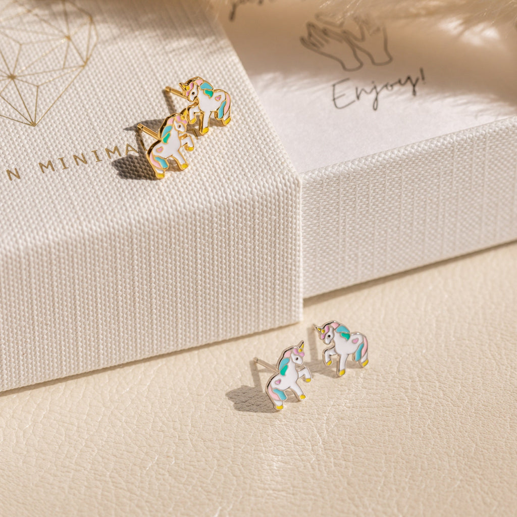 Two pairs of Unicorn Studs—small, colorful pastel enamel earrings—are displayed on a textured surface and white jewelry box, making a delightful gift for girls.