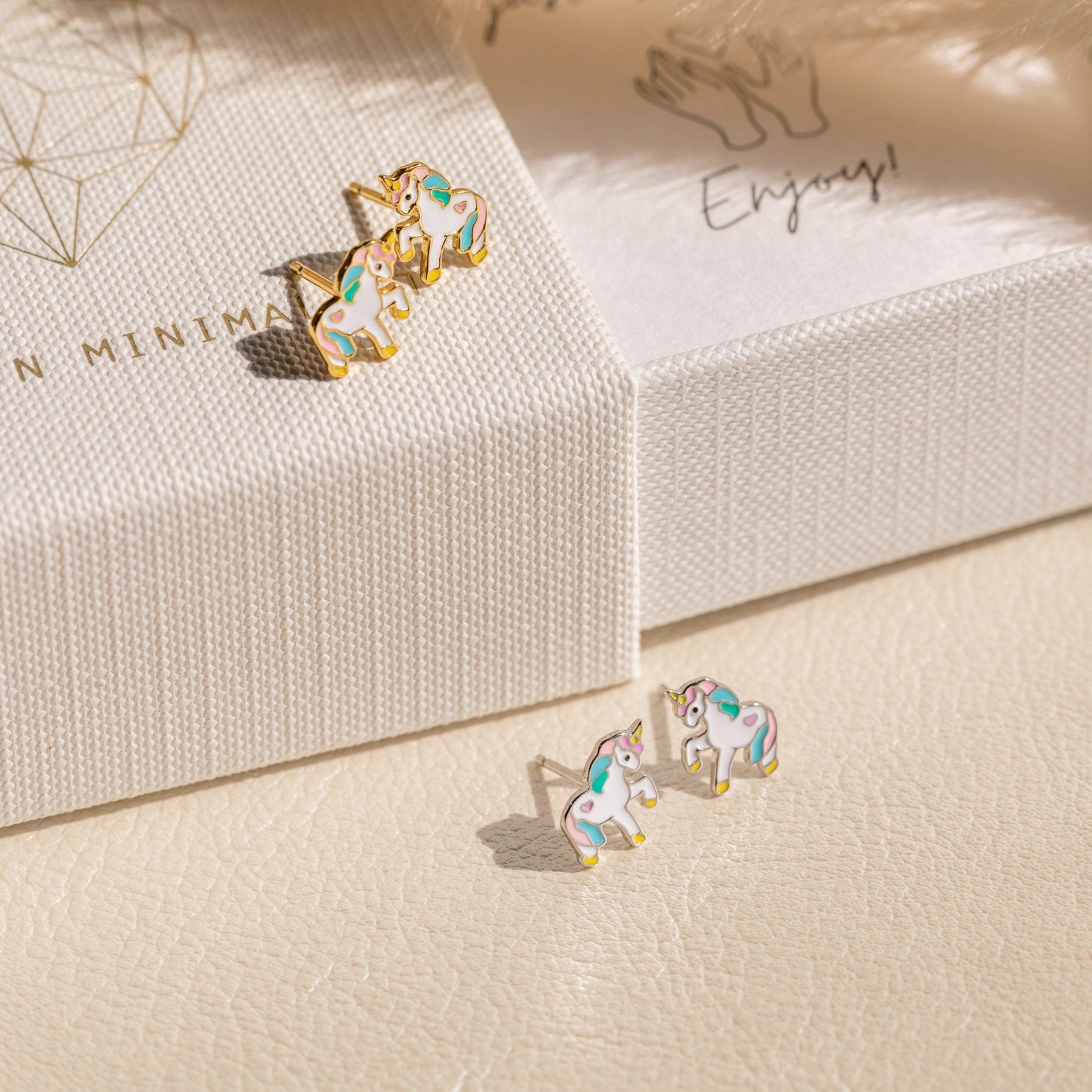 Two pairs of Unicorn Studs—small, colorful pastel enamel earrings—are displayed on a textured surface and white jewelry box, making a delightful gift for girls.