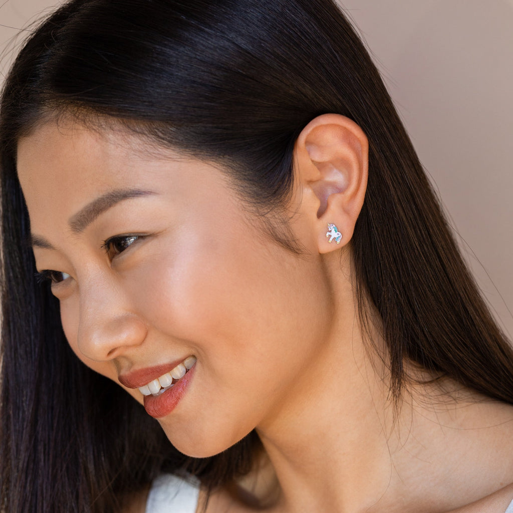 A woman with straight dark hair smiles, wearing a white top and unique Unicorn Studs earrings—a charming gift idea for girls.