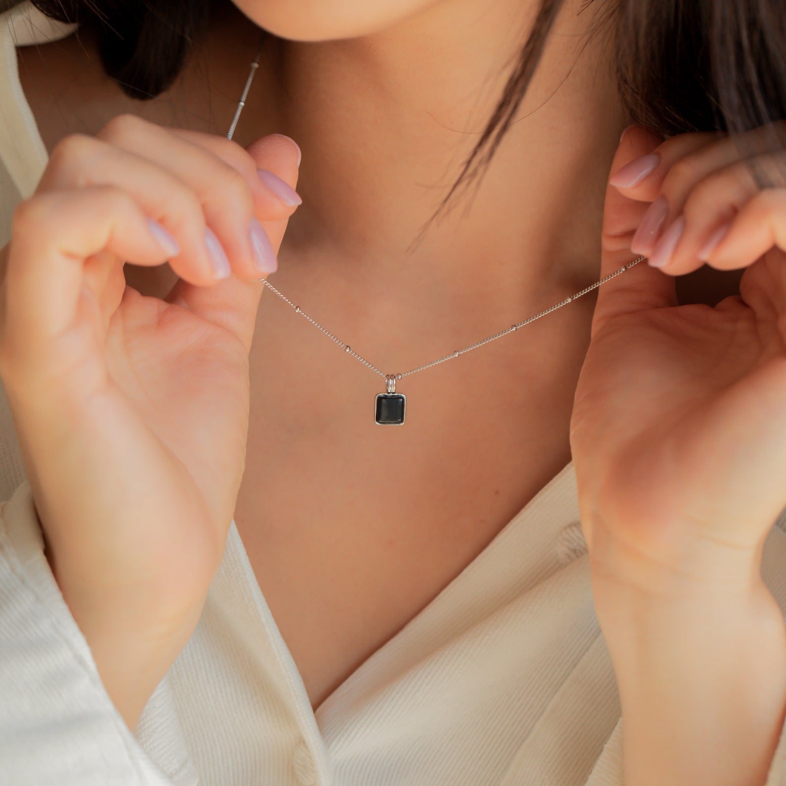 A woman in a white blouse holds the Waverly Black Necklace—a silver satellite chain with a rectangular black enamel pendant—close to her neck.