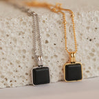 The Waverly Black Necklace features a striking black enamel square pendant on a delicate satellite chain, available in both silver and gold finishes, shown resting on a textured white surface for a modern, elegant look.