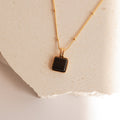 The Waverly Black Necklace features a square black enamel pendant displayed on a textured, light-colored stone surface.