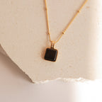 The Waverly Black Necklace features a square black enamel pendant displayed on a textured, light-colored stone surface.