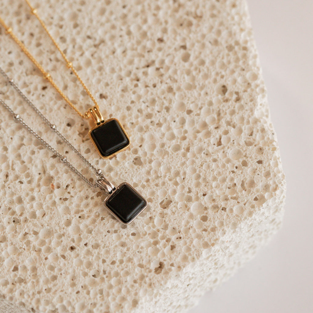 The Waverly Black Necklace, a sleek square pendant on a satellite chain, is displayed on a textured, light-colored stone block.