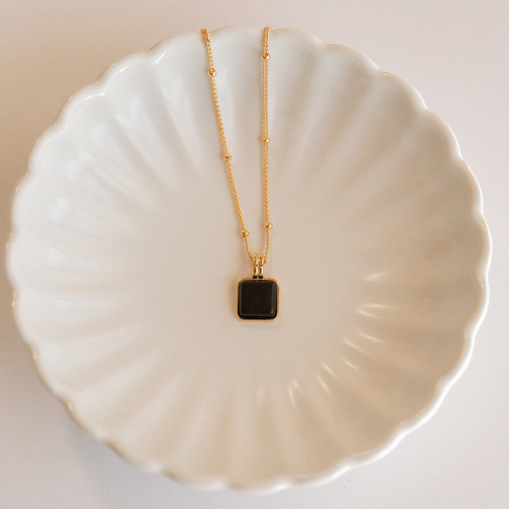 The Waverly Black Necklace, featuring a gold satellite chain and black enamel pendant, is displayed on a white scalloped ceramic dish.