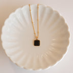 The Waverly Black Necklace, featuring a gold satellite chain and black enamel pendant, is displayed on a white scalloped ceramic dish.