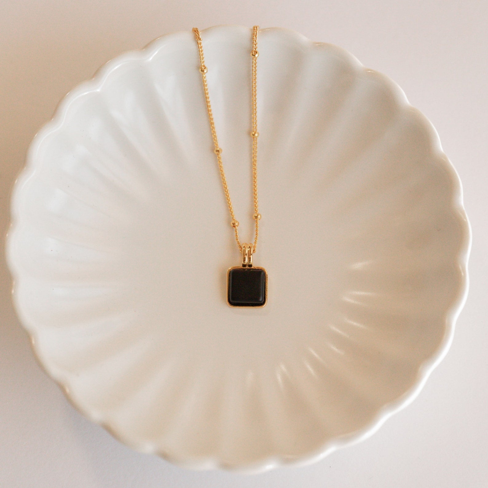 The Waverly Black Necklace, featuring a gold satellite chain and black enamel pendant, is displayed on a white scalloped ceramic dish.