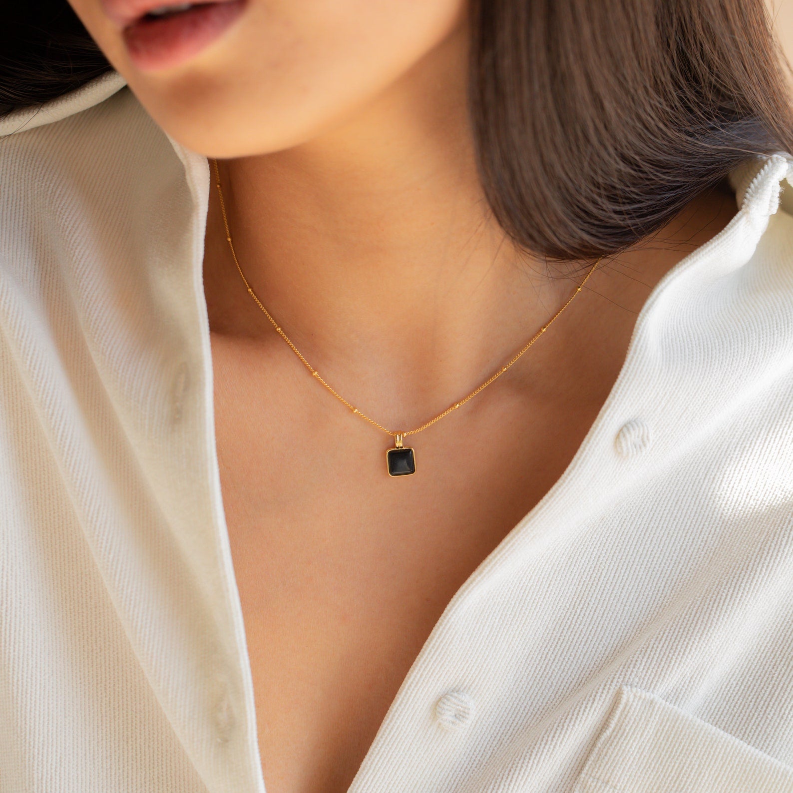 A woman in a white shirt wears the Waverly Black Necklace, a gold satellite chain adorned with a small black enamel pendant.