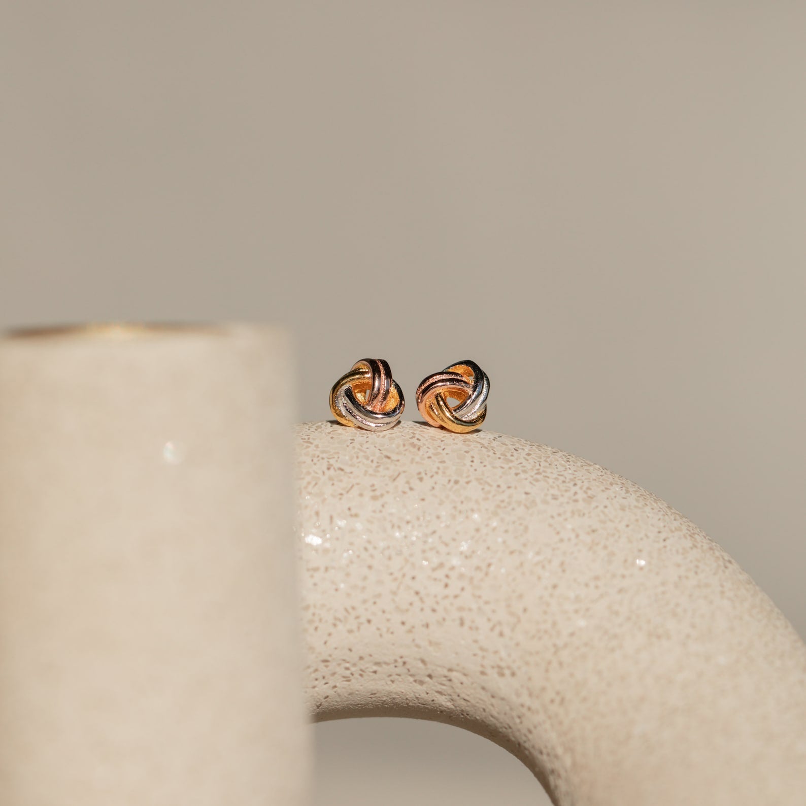 The Trinity Knot Studs, featuring a delicate twisted design, are displayed on a curved, textured beige ceramic surface with soft lighting—making them an elegant option for an anniversary gift.