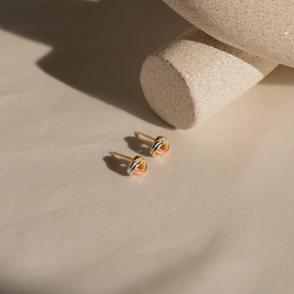 Trinity Knot Studs rest on cream fabric next to a speckled beige ceramic piece in sunlight—timeless earrings that make an ideal anniversary gift.