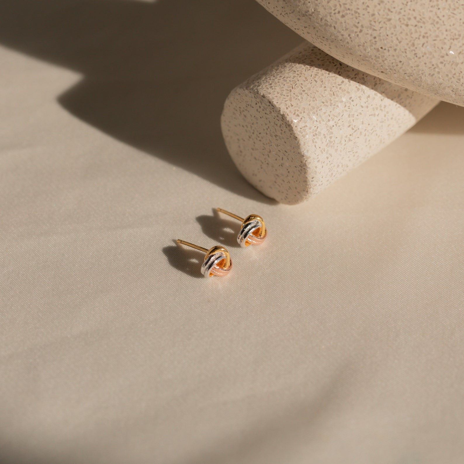 Trinity Knot Studs rest on cream fabric next to a speckled beige ceramic piece in sunlight—timeless earrings that make an ideal anniversary gift.