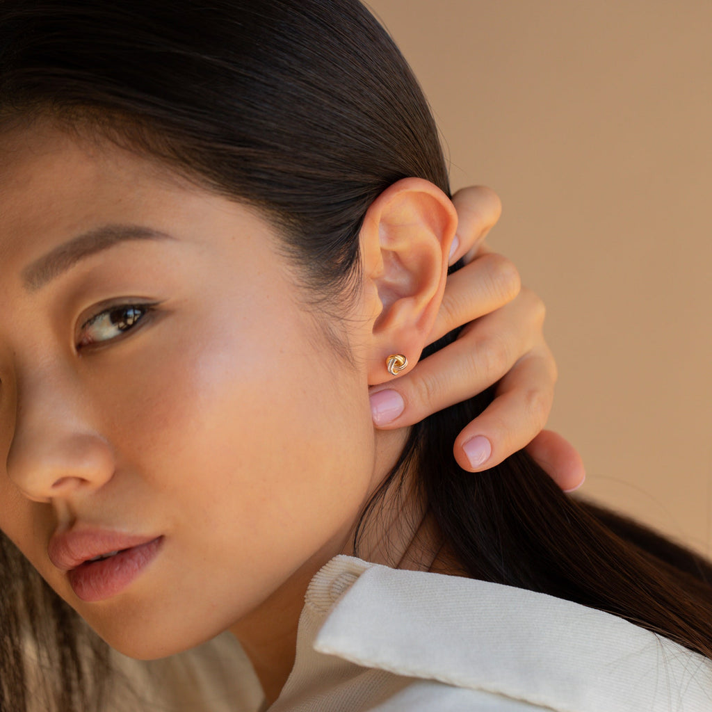 A woman with long dark hair touches her ear, showcasing Trinity Knot Studs and a cream collared shirt—an ideal anniversary gift.