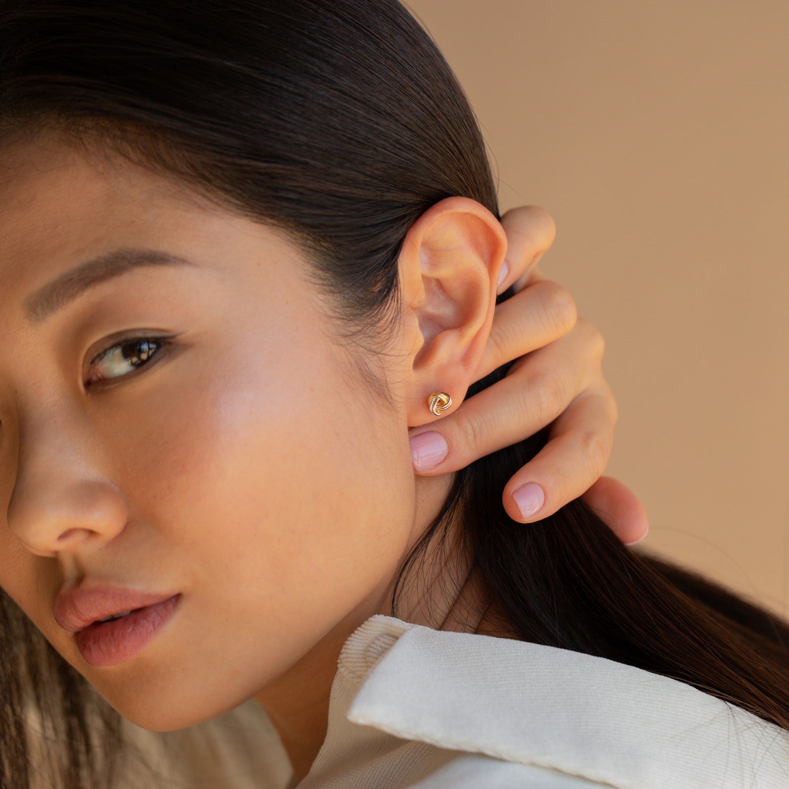 A woman with long dark hair touches her ear, showcasing Trinity Knot Studs and a cream collared shirt—an ideal anniversary gift.